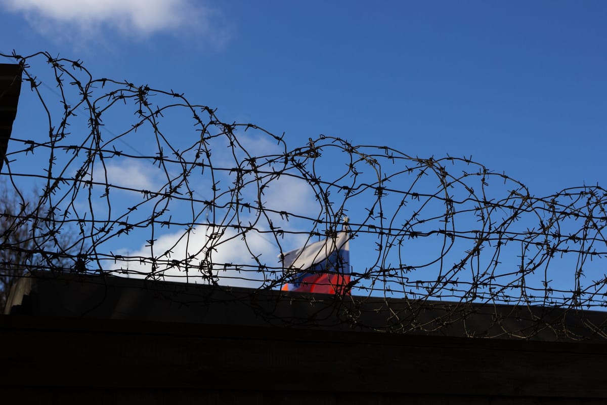 A Russian flag flutters behind a coiled barbed wire security fence under a blue sky.
