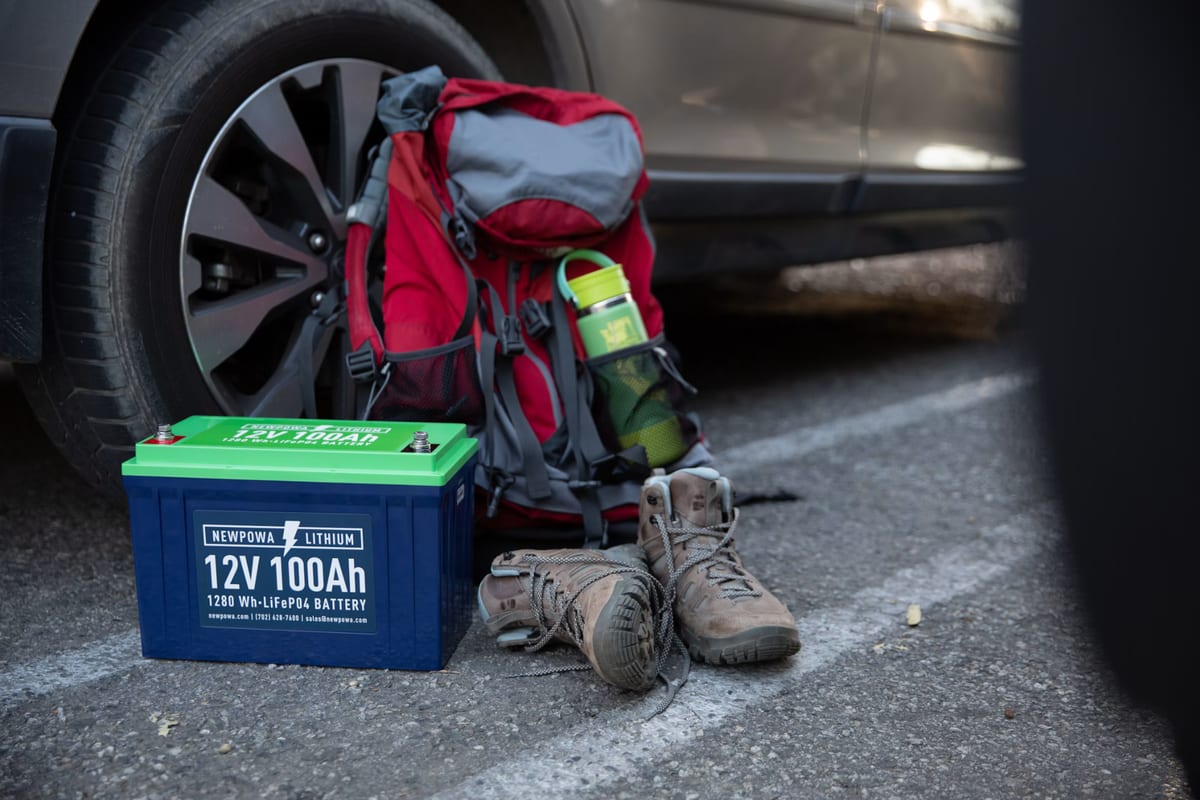 A lithium battery, red backpack, and hiking boots placed beside a car tire on the pavement.