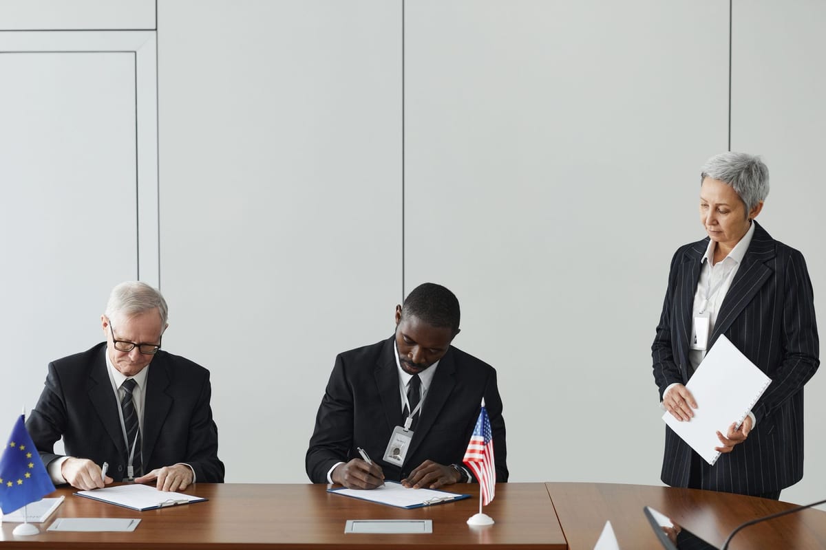 Diplomats signing an international agreement at a conference table with European Union and United States flags.