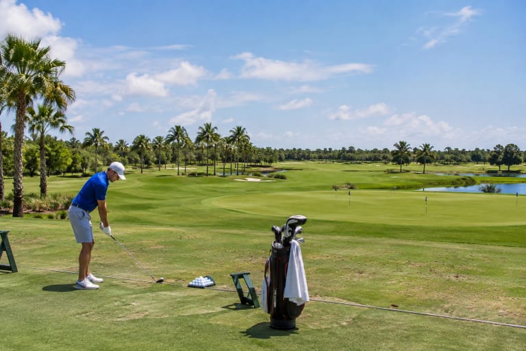 A golfer practices his swing on a sunny Florida driving range with palm trees and a golf bag nearby.