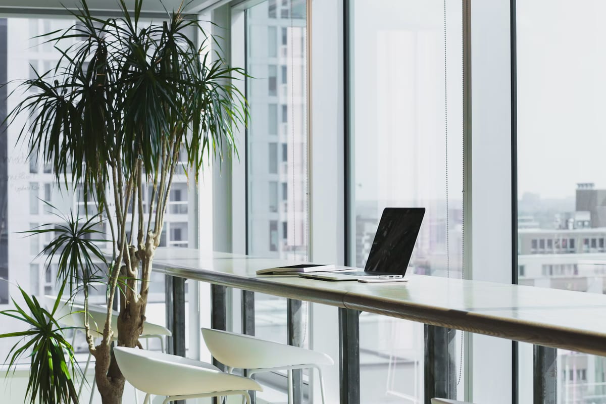 A laptop on a high desk beside large office windows with bright natural daylight illuminating the workspace.