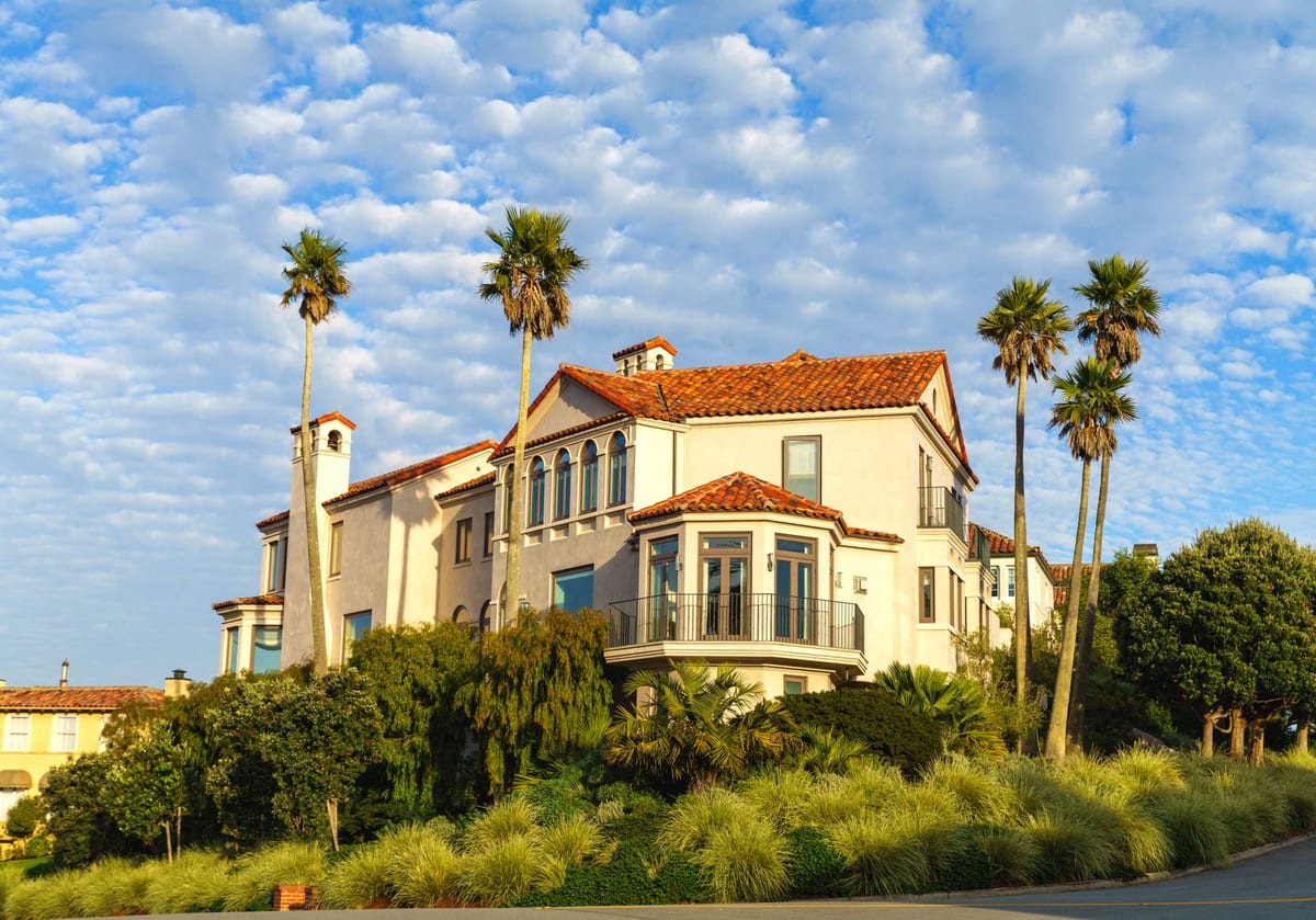 A large Mediterranean-style luxury estate with a red tile roof and palm trees under a partly cloudy sky.