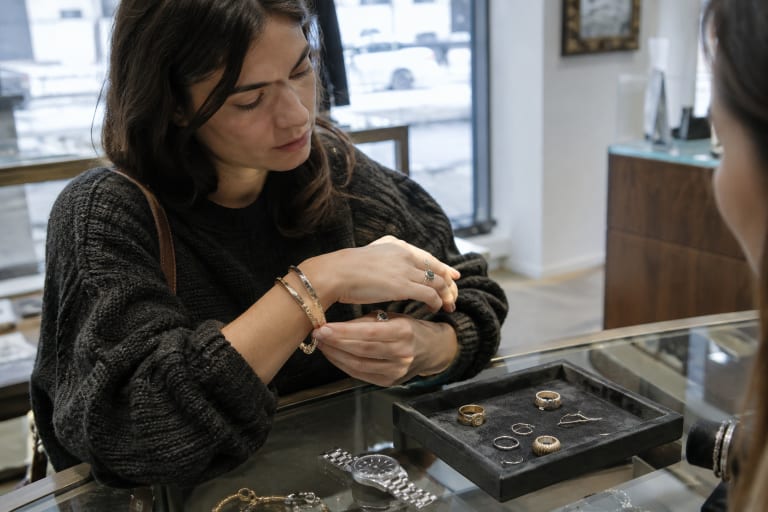 Woman trying on gold bracelets and rings over a glass counter in a jewelry store.